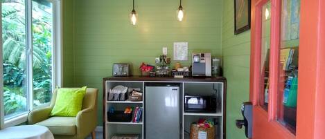 A compact kitchen with a refrigerator, microwave, toaster, and coffee maker. A small seating area with a chair and table is adjacent to the kitchen.