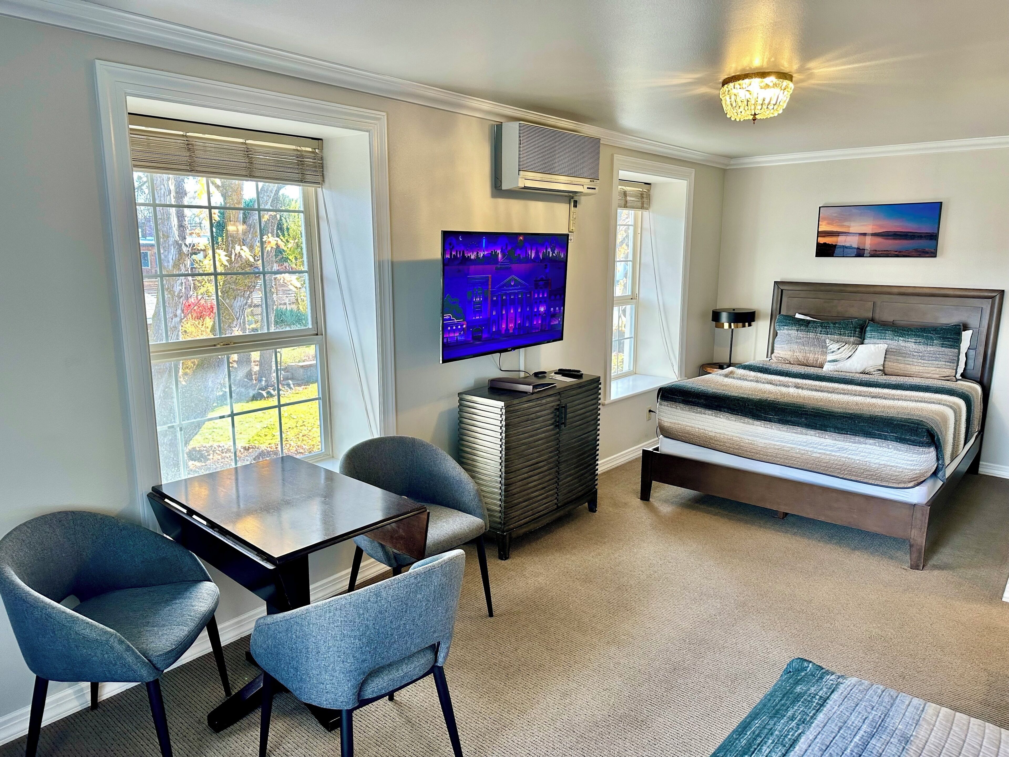 A bedroom with a bed, a TV, a desk with chairs, and a window.