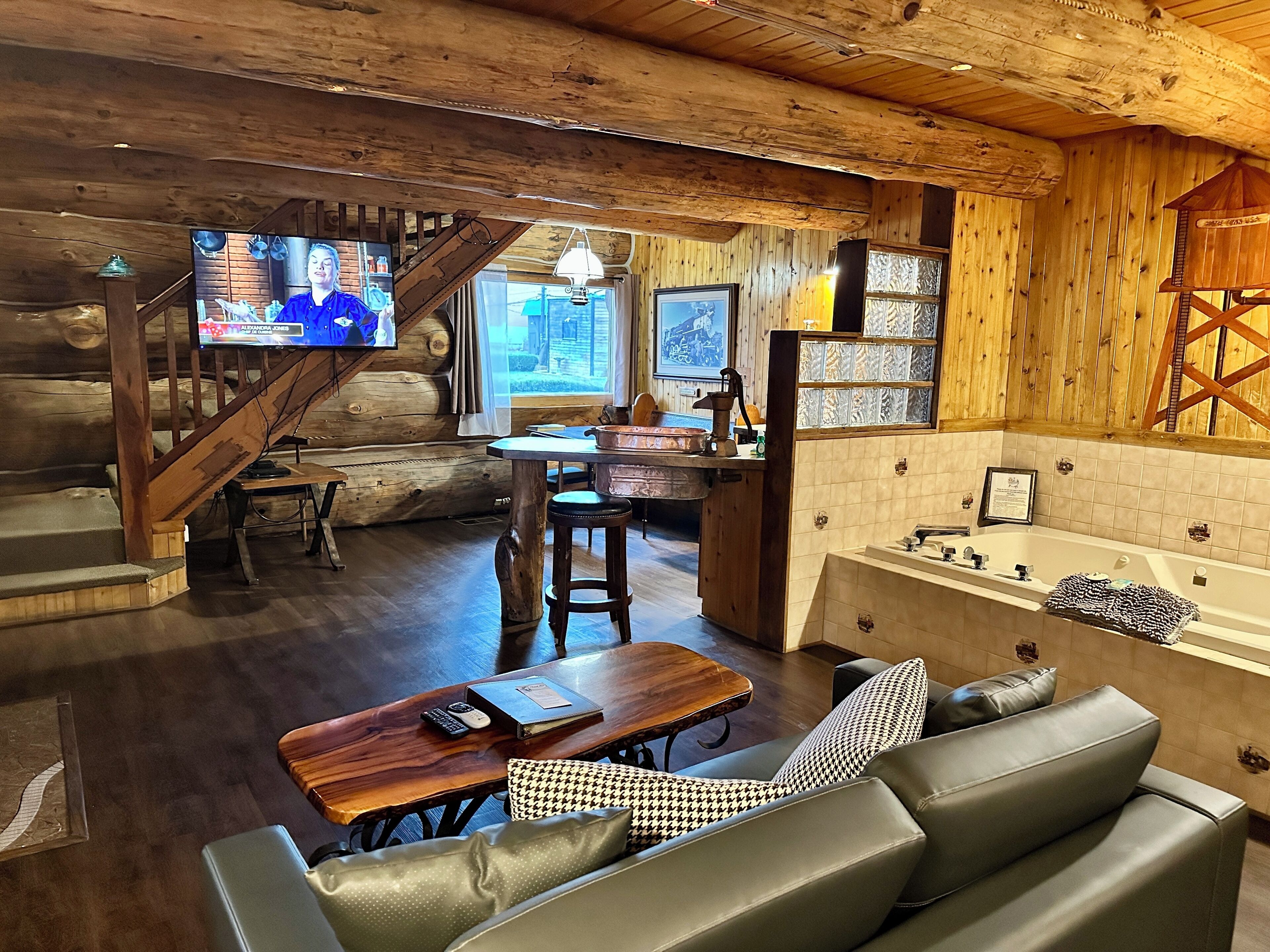 A log cabin interior with a wooden staircase, a dining area, a kitchen with a sink and stove, and a living room with a sofa and a television.