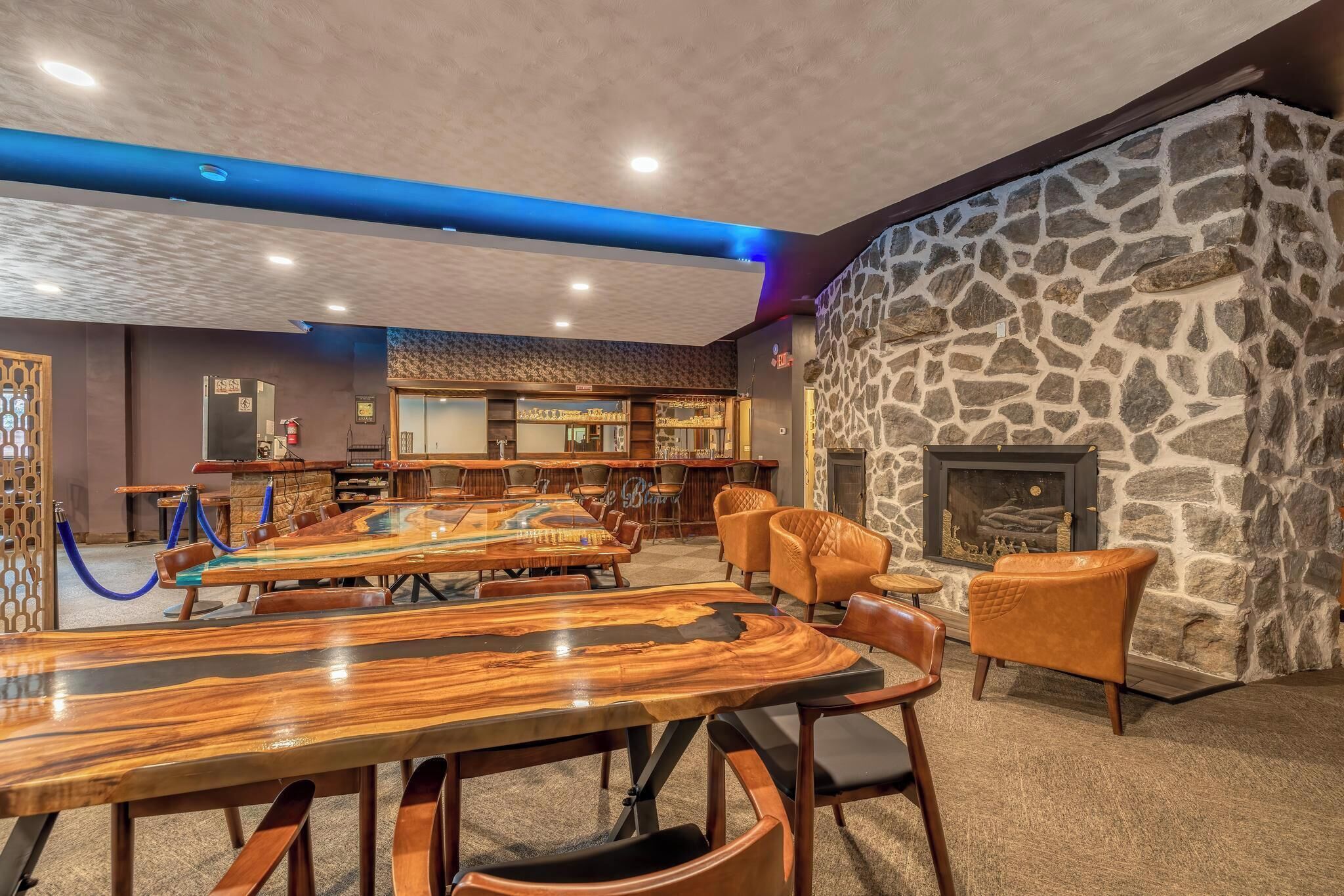 A spacious room with a large wooden table and chairs, a stone wall with a fireplace, and a bar area in the background.