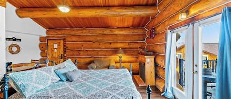 A log cabin bedroom with a bed, bedside table, lamp, and a view through a door.