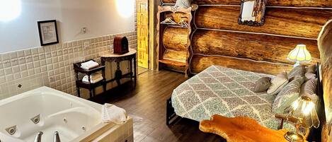 Un dormitori d'estil cabana de fusta, amb un llit gran de fusta, una nitzeta de fusta amb una làmpada, una cadira de fusta, una taula petita amb una làmpada i un bany amb banyera i barra de tovalloles.