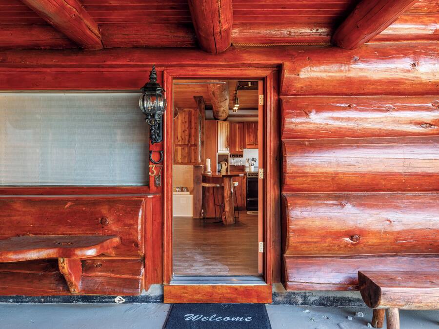 A wooden cabin with a wooden bench outside, a welcome mat, and a door leading to a kitchen area.