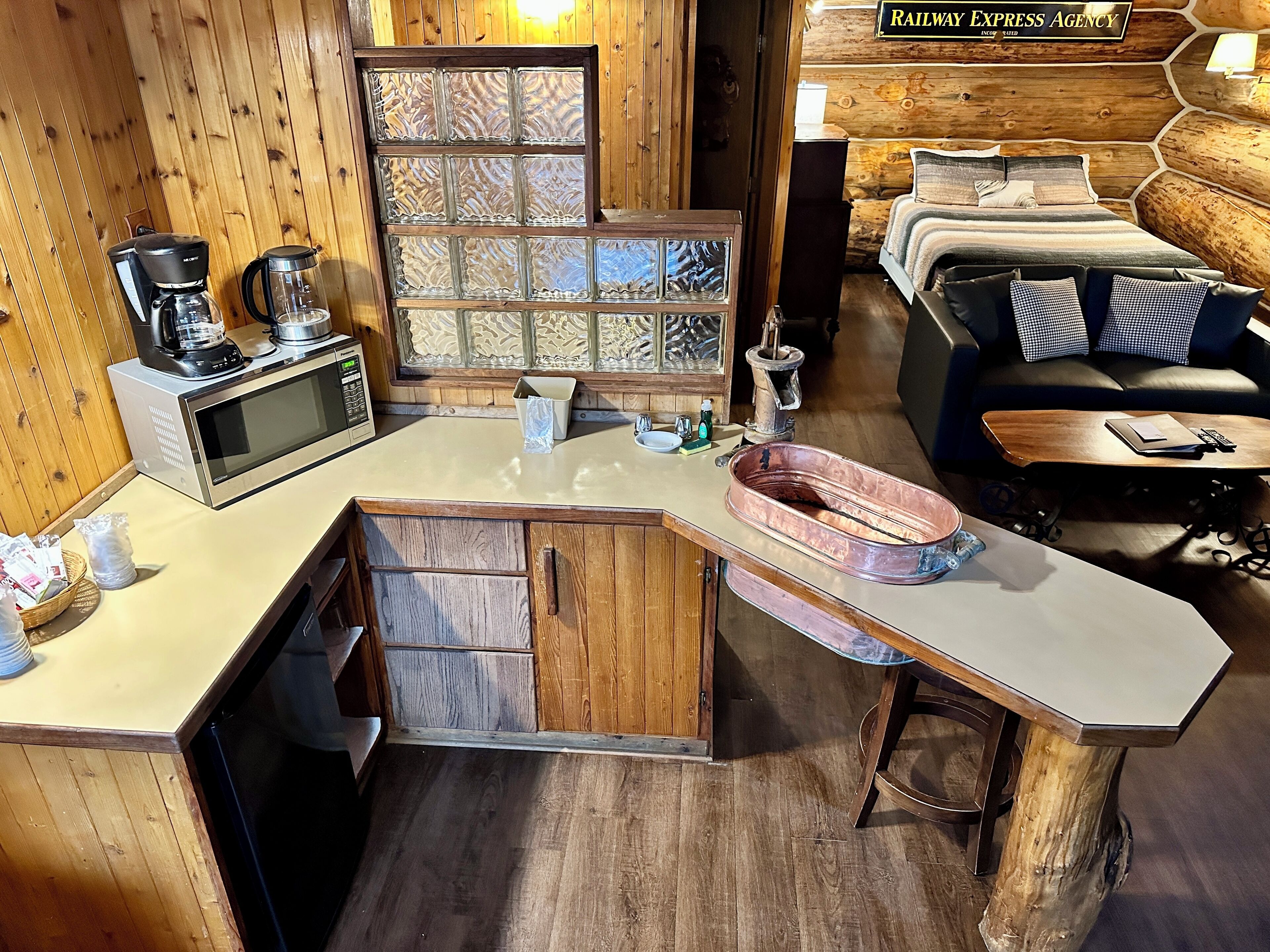 A compact kitchen with a microwave, coffee maker, and kettle. There's a wooden island with a sink and a small seating area. A bed with checkered pillows is visible in the background.