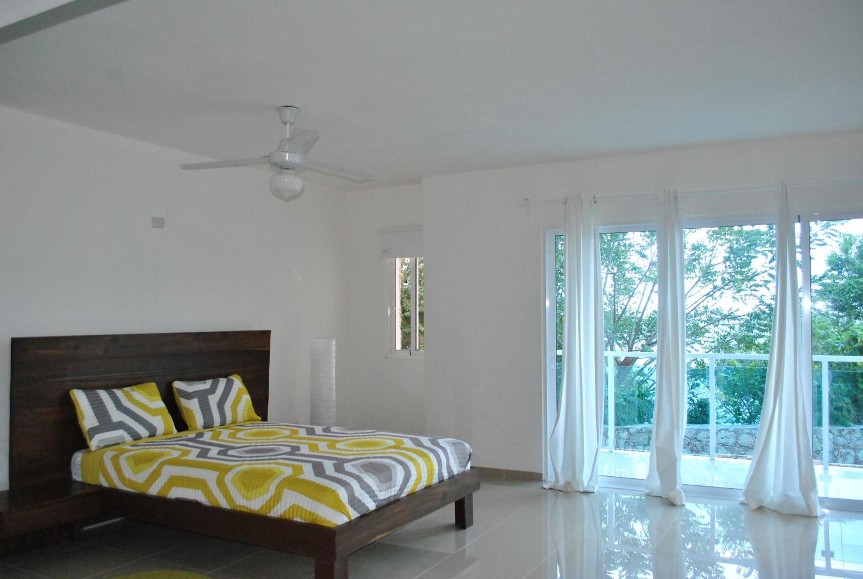 A bedroom with a bed, a ceiling fan, and a view of the outdoors through large windows.