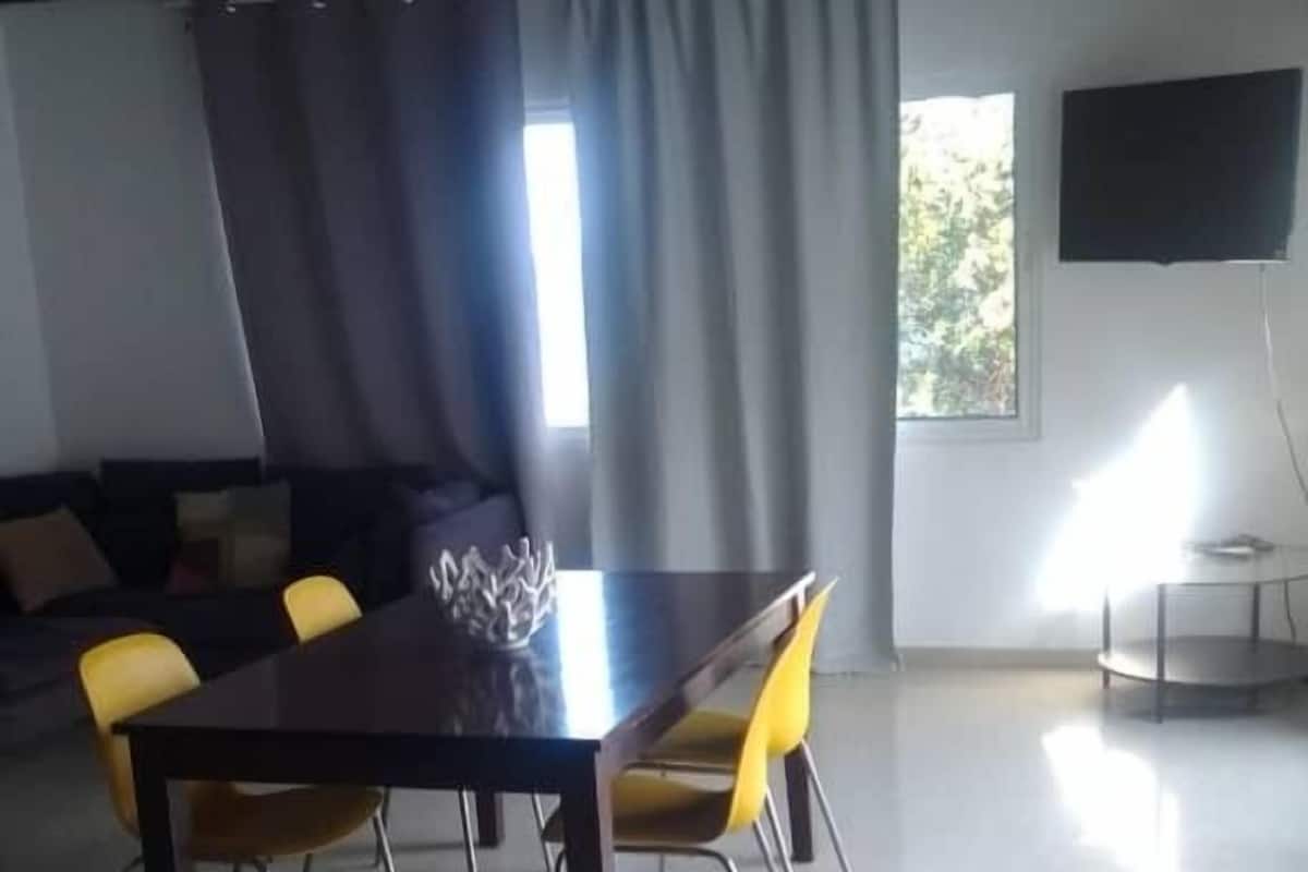 Un coin repas comprenant une table et des chaises, un canapé, un téléviseur et une fenêtre avec des rideaux.