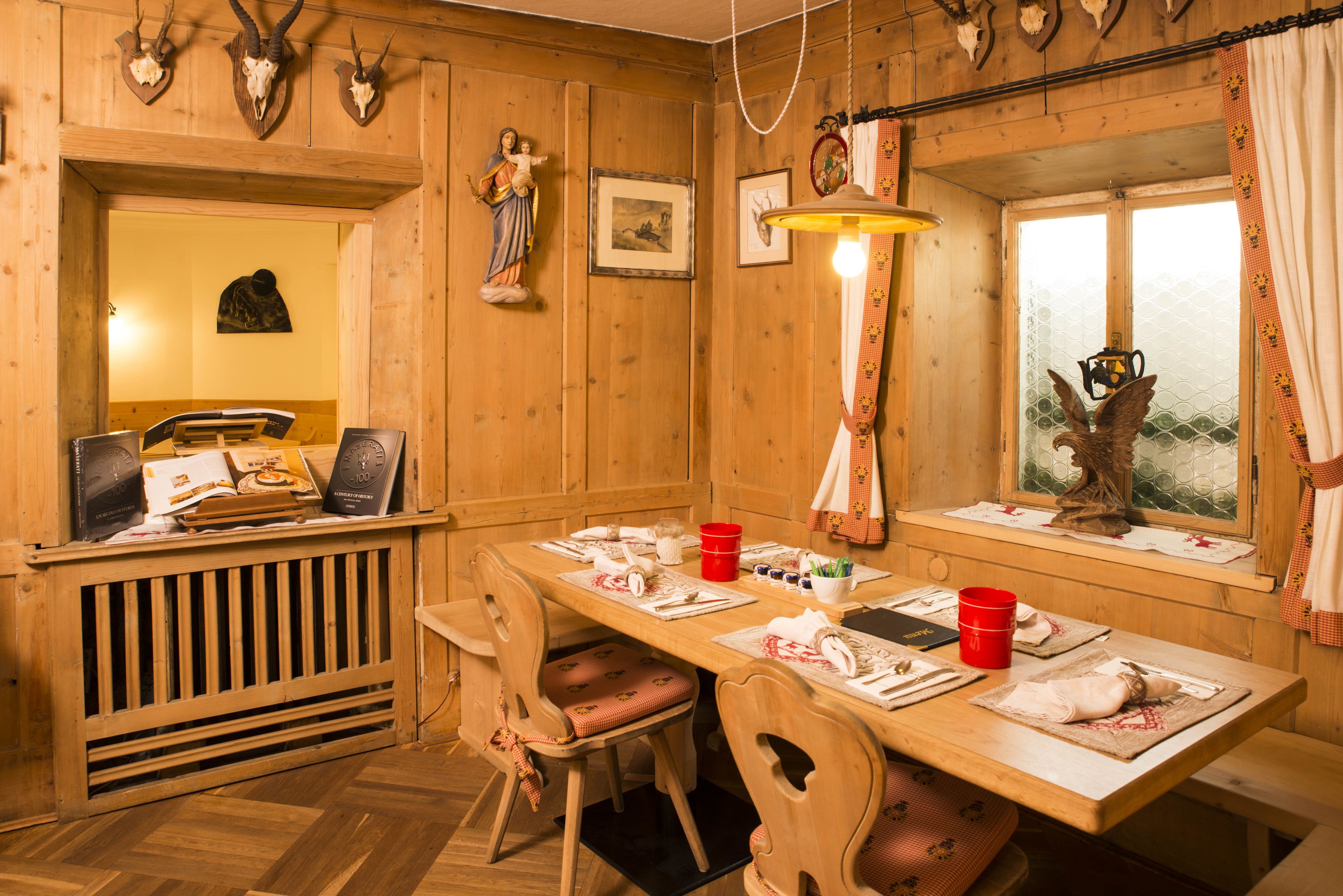A wooden dining room with a long table set for four, chairs, a red bucket, and a window with a decorative bird.
