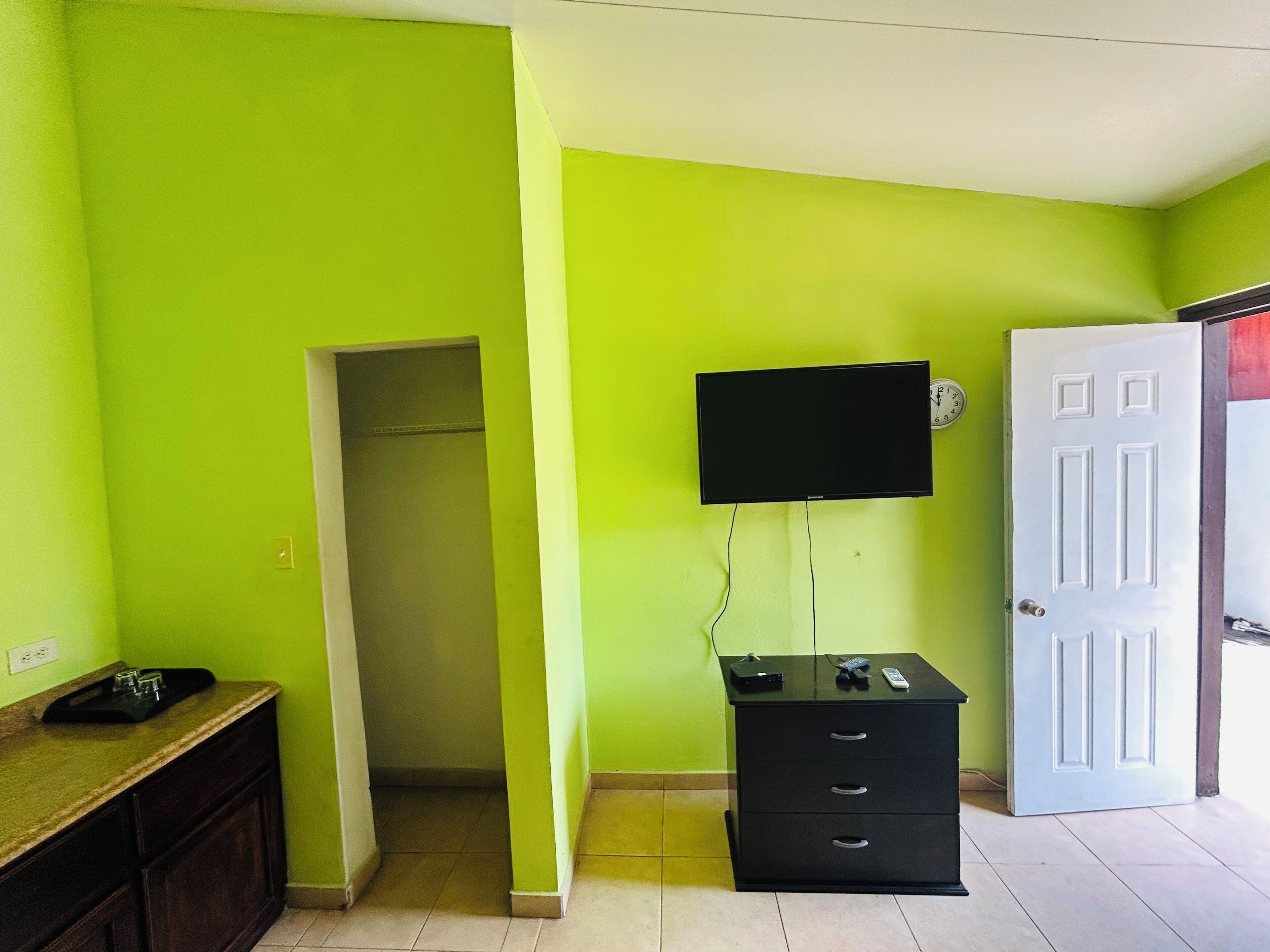 A room with a green wall, a white door, a black TV mounted on the wall, a black filing cabinet, and a wooden cabinet with a countertop.