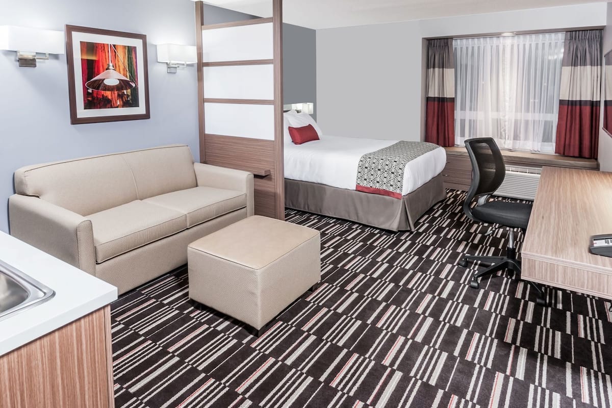 Ein Hotelzimmer mit Bett, Sofa, Schreibtisch, Stuhl, kleinem Tisch, Spüle und einem Fenster mit Vorhängen.
