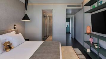 Una habitación de hotel moderna con una cama grande, una lámpara de noche, un televisor sobre una estantería y un vestidor con ducha.