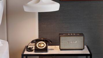Una habitación de hotel moderna con una cama blanca, una mesita de noche con un teléfono antiguo y un altavoz Marshall.