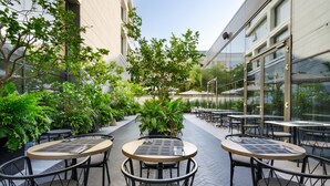 An outdoor patio area with round tables and chairs, surrounded by lush greenery and a building with large windows.
