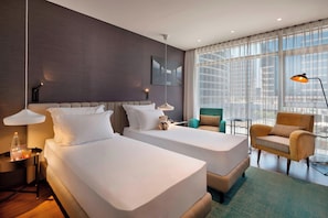 Habitación de hotel con dos camas, un sofá, una silla, una mesita y un ventanal con vistas a la ciudad.