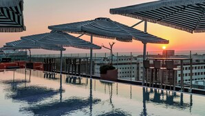 A rooftop infinity pool with striped umbrellas and seating areas, overlooking a cityscape at sunset.