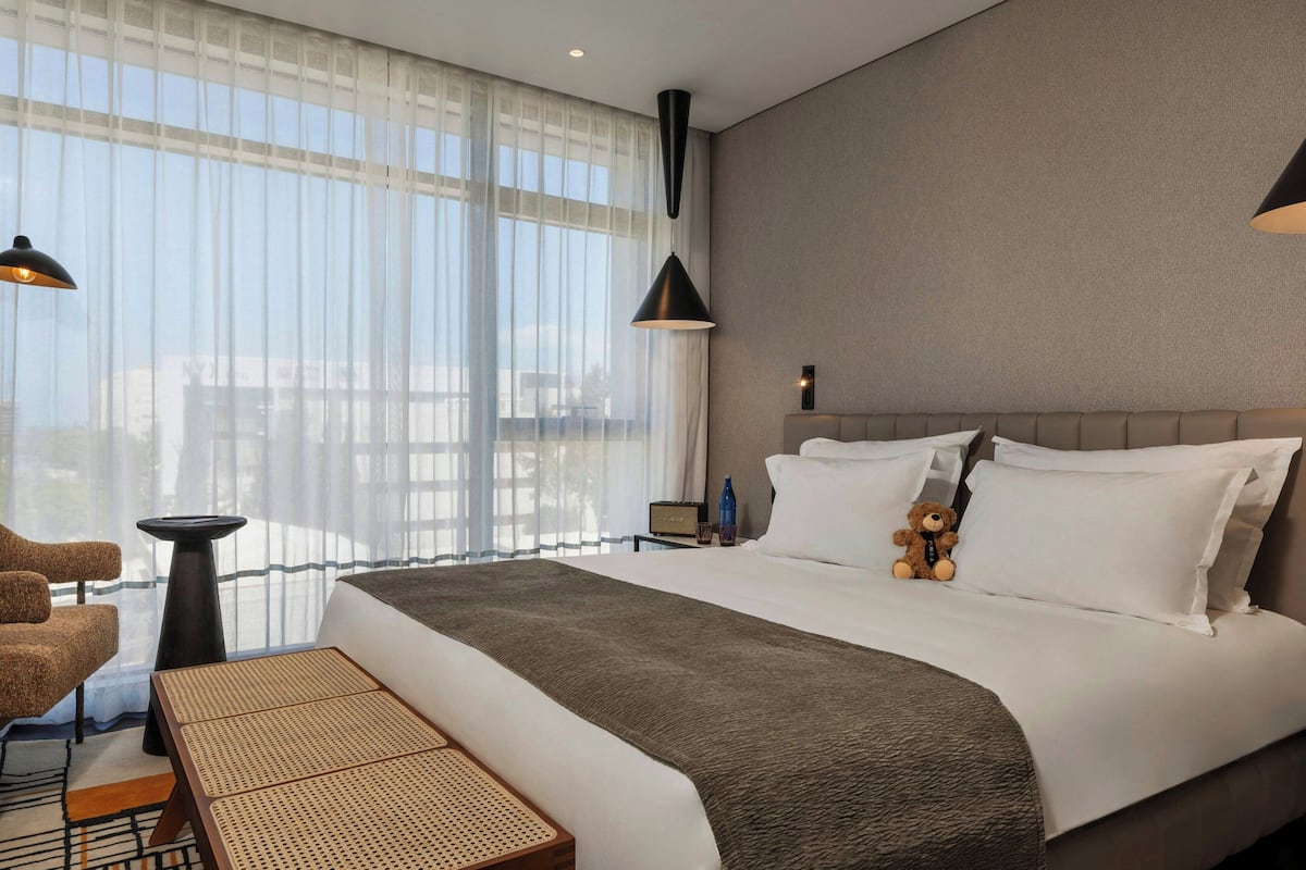 A hotel room with a large bed, a chair, a small table, and a view of the city through sheer curtains.