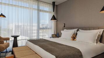 Camera d'albergo con un letto grande, una sedia, un tavolino e vista sulla città attraverso tende leggere.