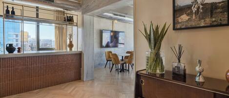 Un interior moderno con suelo de espiga, un mostrador de recepción de madera, una zona de comedor con sillas, un ventanal grande con botellas en el alféizar, y un cuadro en la pared.