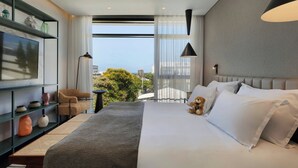 A modern hotel room with a large bed, a television on a shelf, a view of the city, and a teddy bear on the bed.