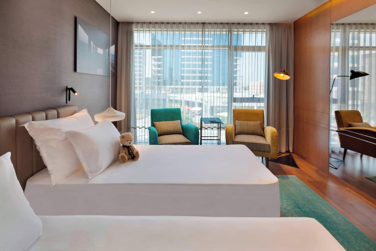 A hotel room with a large bed, two armchairs, a small table, and a view of the city through large windows.