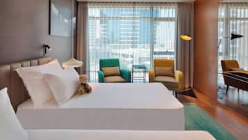 A hotel room with a large bed, two armchairs, a small table, and a view of the city through large windows.