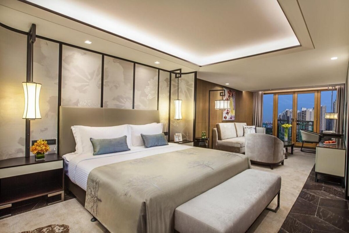 A modern hotel room with a large bed, bedside lamps, a seating area with a chair, and a view of the cityscape through large windows.