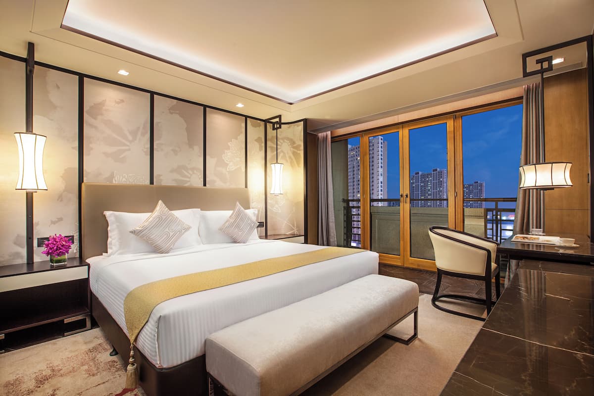 Una habitación de hotel con una cama grande, un banco, un escritorio y vista a la ciudad de noche.