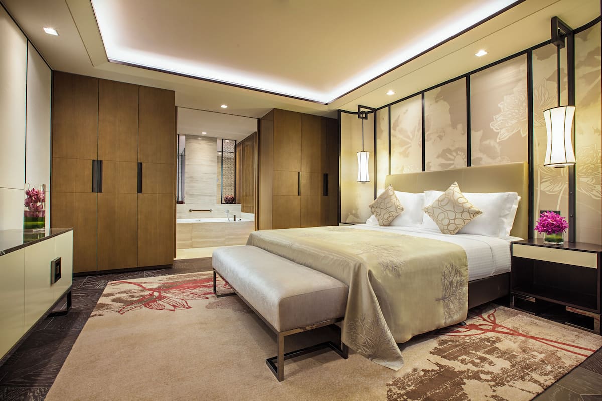 Una habitación de hotel moderna con una cama grande, un banco, una mesita de noche y un baño visible a través de una puerta abierta.