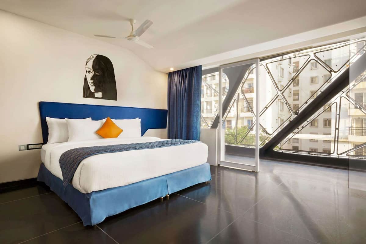 A modern hotel room with a large bed, a ceiling fan, and a view of the cityscape through a large window.