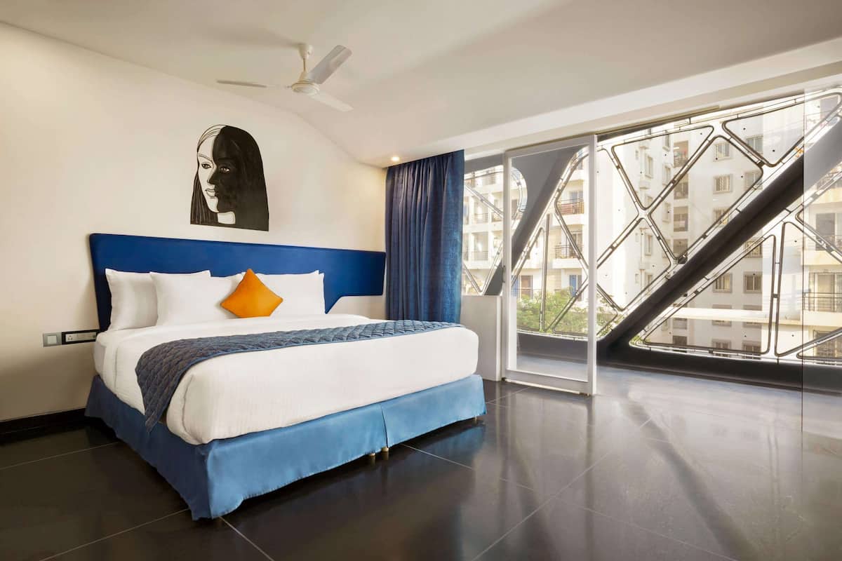 A modern hotel room with a large bed, a ceiling fan, and a view of the city through a large window.
