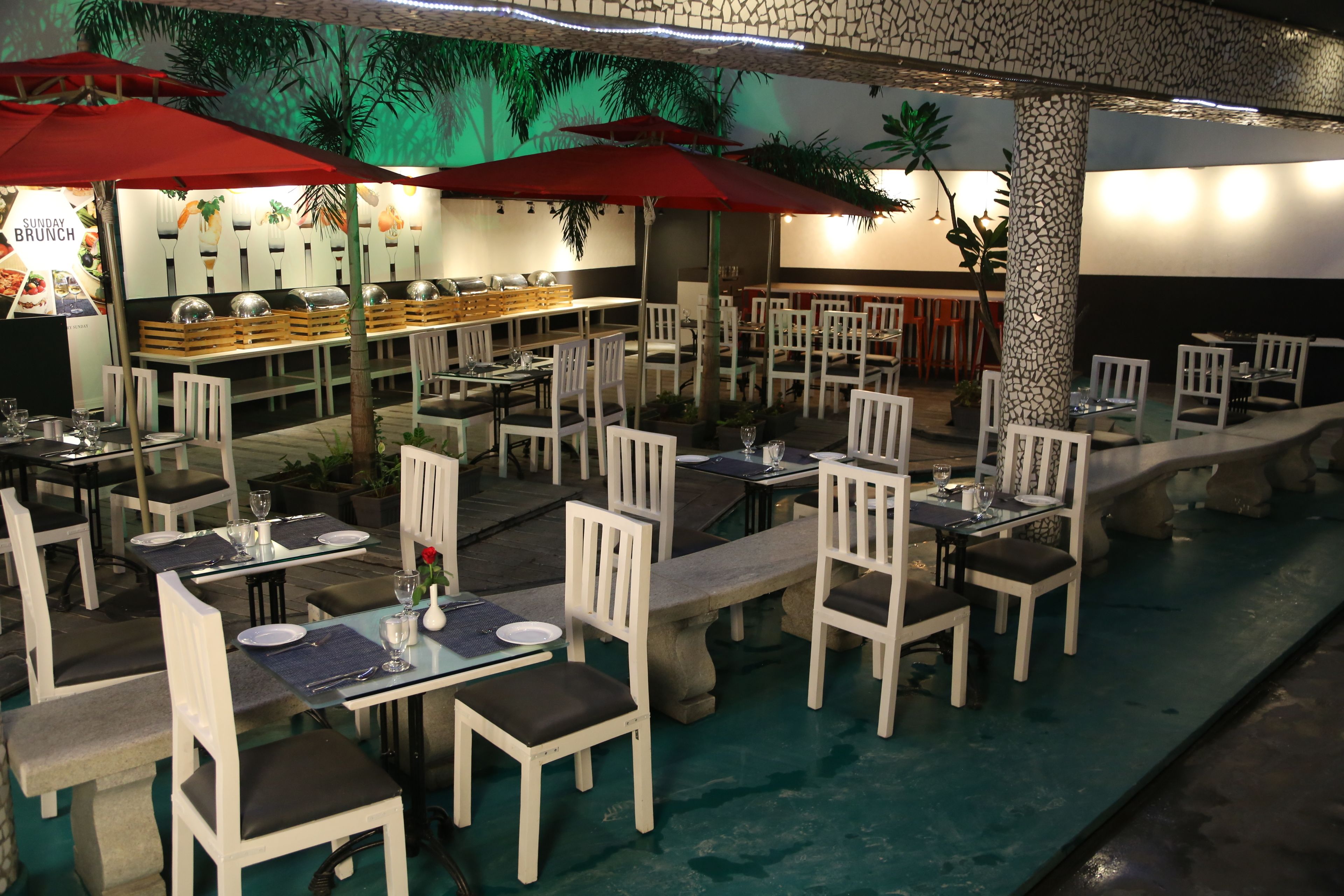 An outdoor dining area with white chairs and tables, red umbrellas, and a pool.