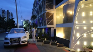 Un edificio moderno con una fachada geométrica distintiva, una alfombra roja que conduce a la entrada y un coche blanco estacionado al frente.