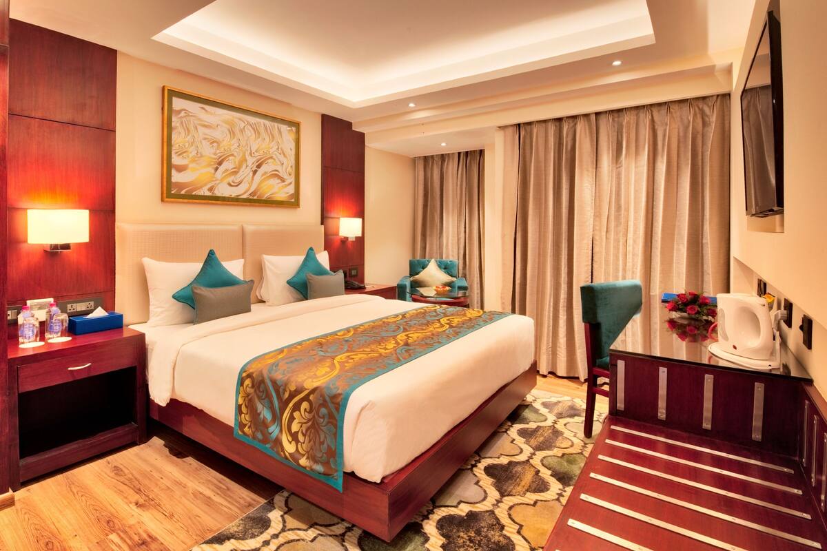 A hotel room with a large bed, a desk with a chair, a nightstand, a wall-mounted TV, and a painting.
