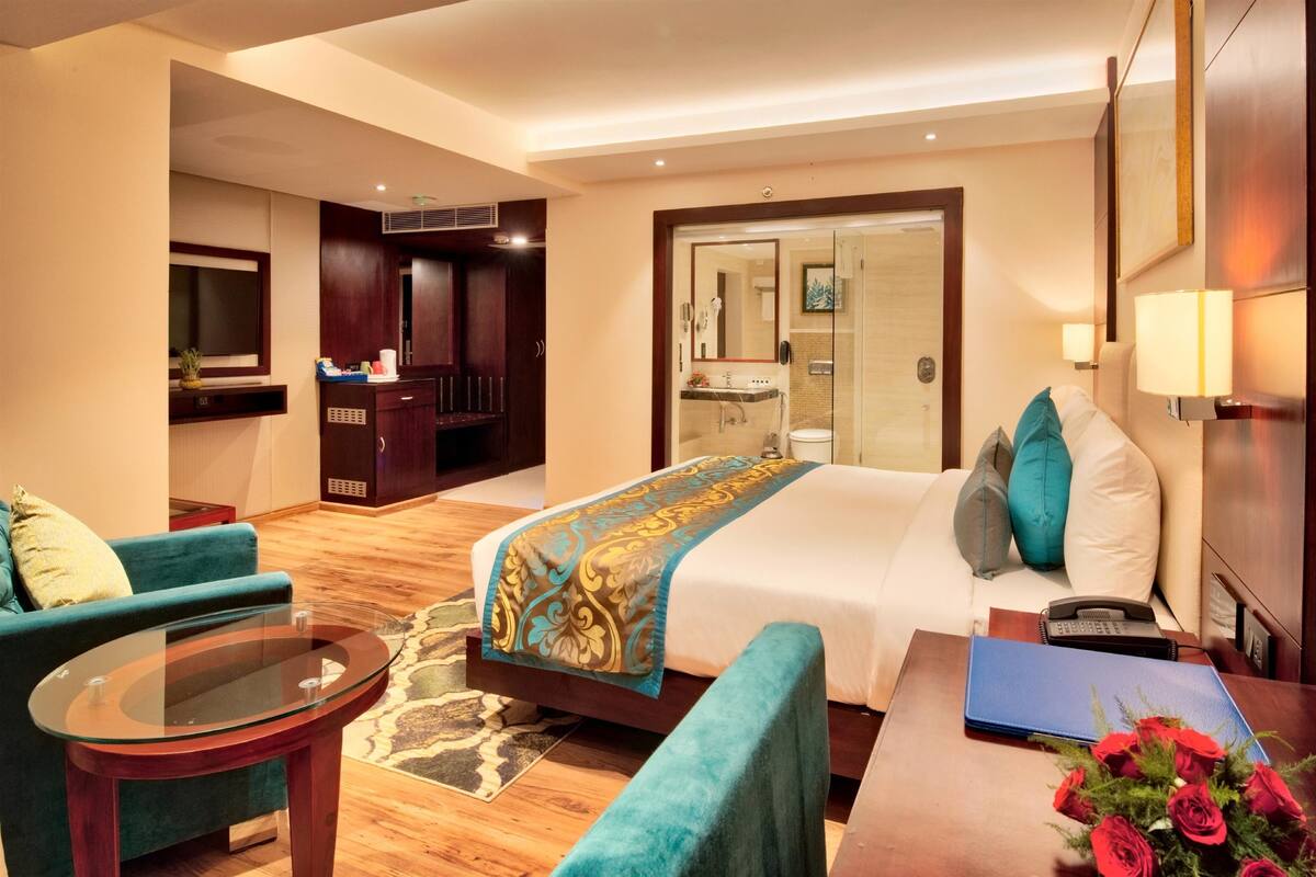 A hotel room with a bed, a sofa, a glass table, a desk with a laptop and flowers, a TV, a wardrobe, and a bathroom visible through an open door.