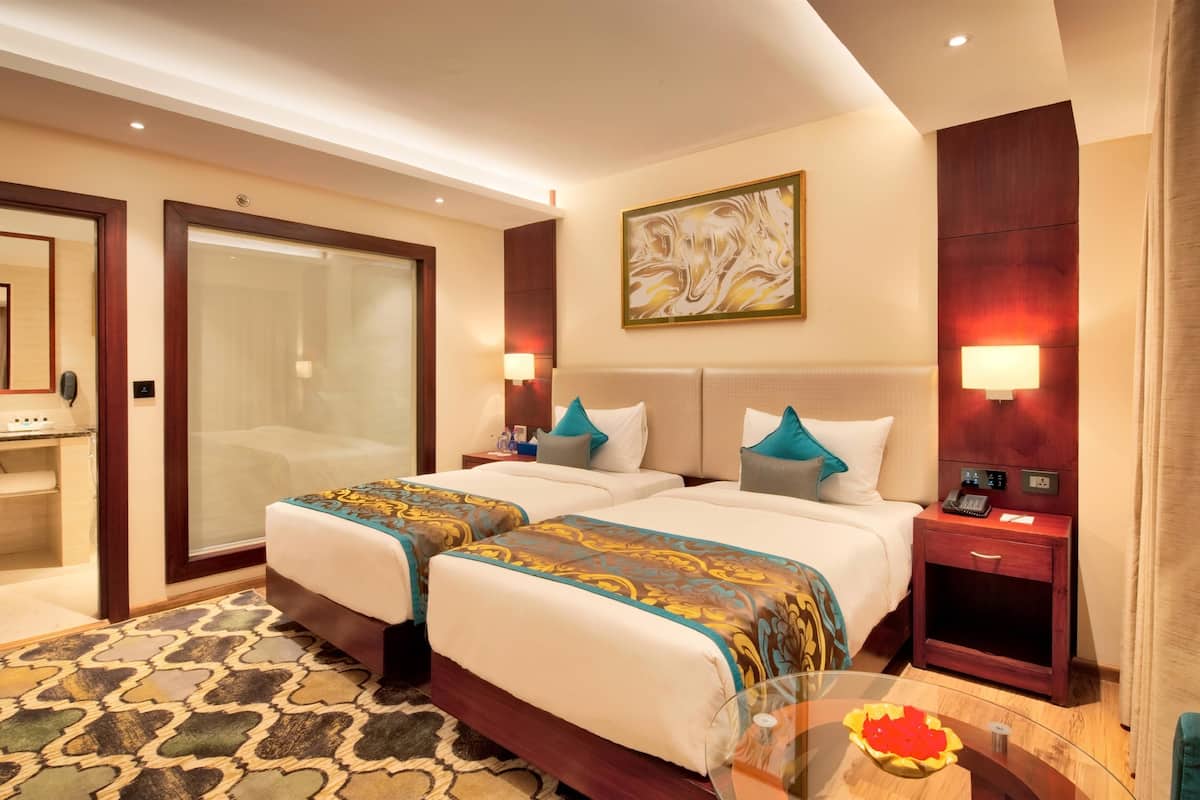 A hotel room with two beds, a glass shower enclosure, a bedside table with a lamp, and a painting on the wall.