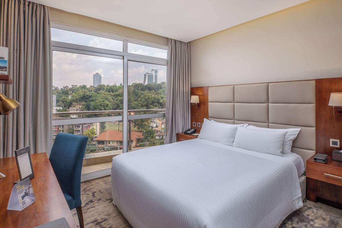 Hotelkamer met een groot bed, een bureau met stoel, een nachtkastje en een stadszicht door grote ramen.