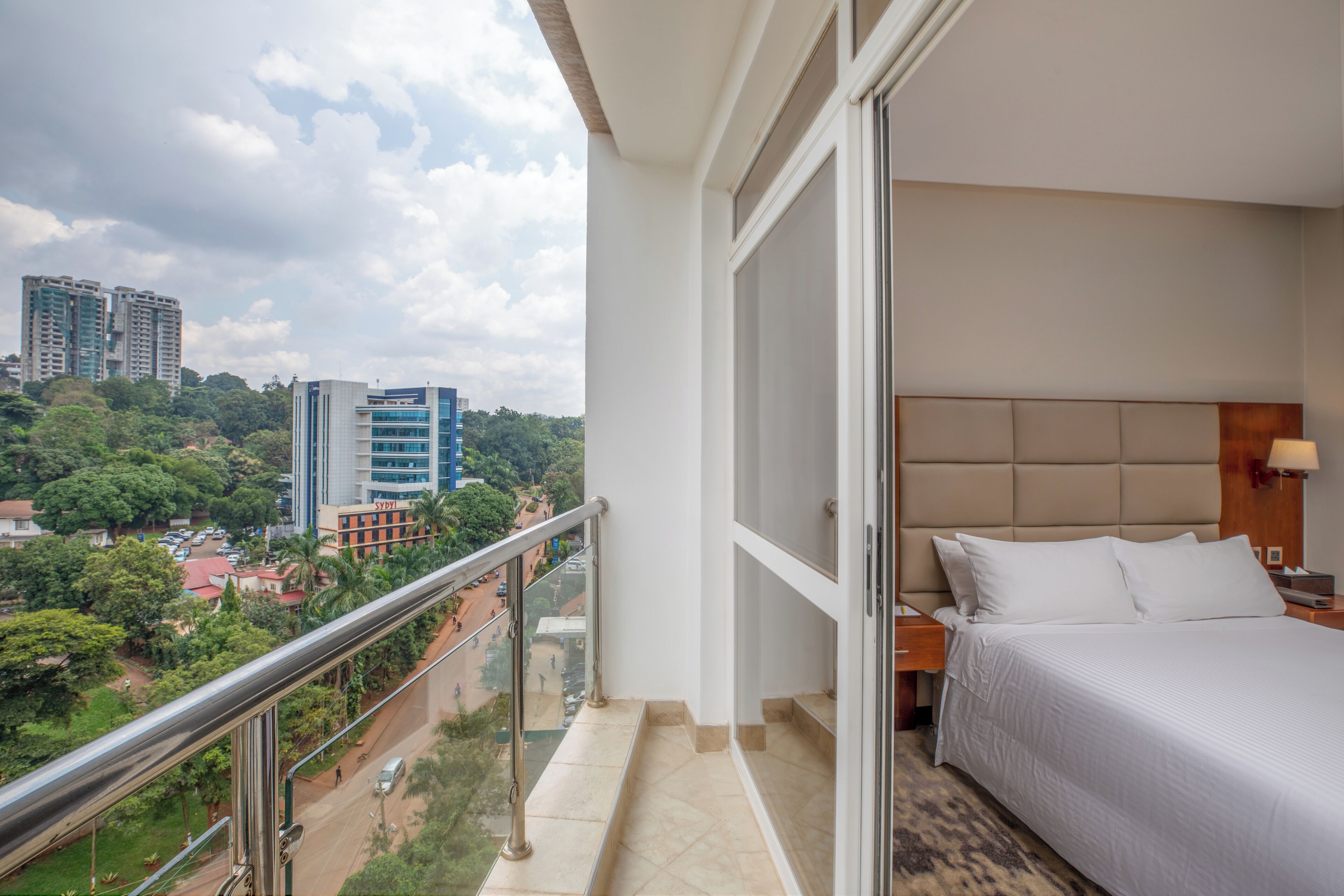 Ein Hotelzimmer mit Balkon und Blick auf eine Stadtlandschaft, ein Bett mit weißer Bettwäsche sowie eine Aussicht auf hohe Gebäude und Grünflächen.