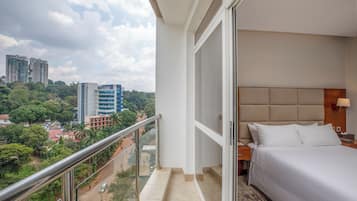 Ein Hotelzimmer mit Balkon und Blick auf eine Stadtlandschaft, ein Bett mit weißer Bettwäsche sowie eine Aussicht auf hohe Gebäude und Grünflächen.