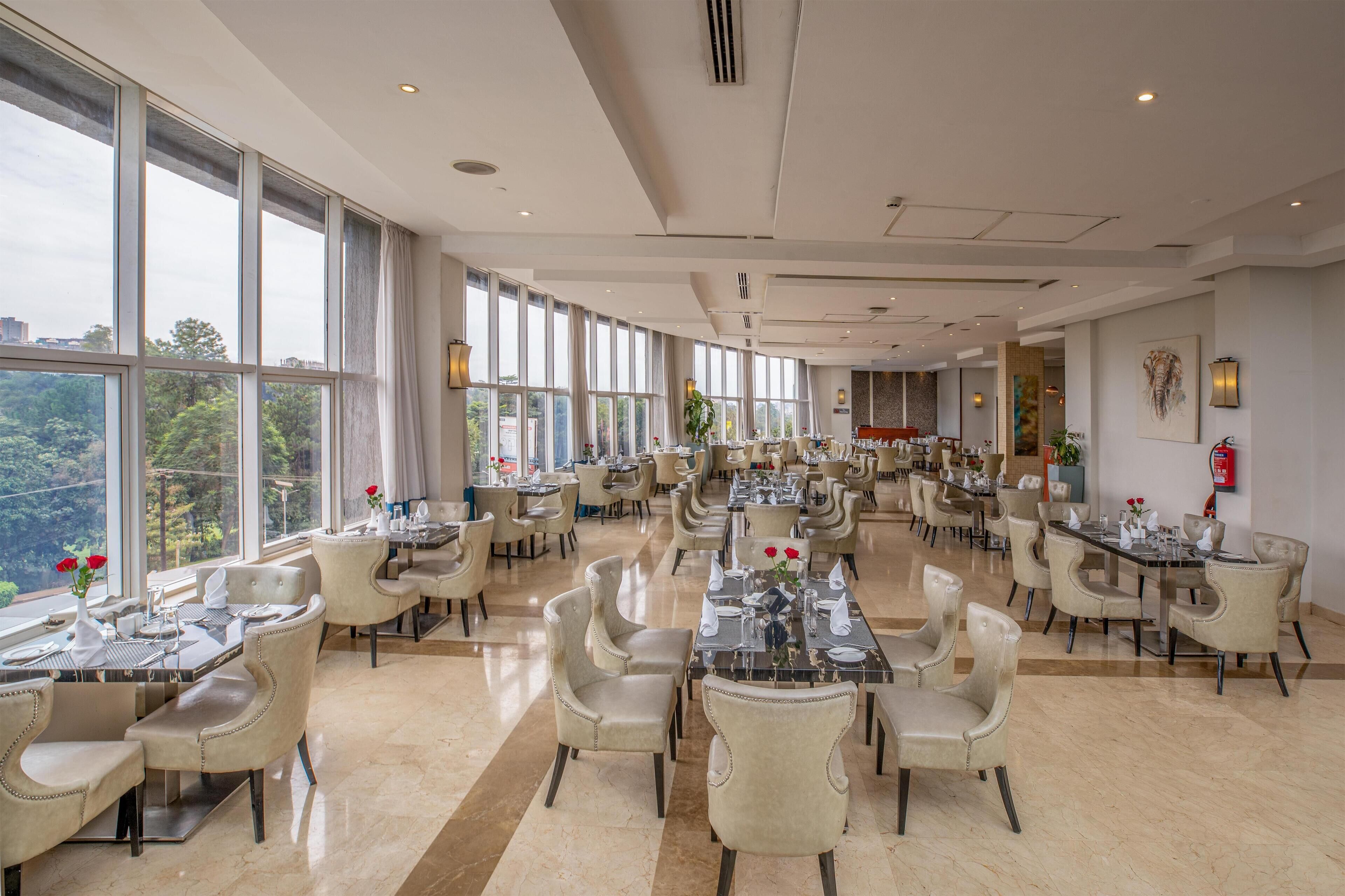 A spacious dining area with multiple tables and chairs, large windows offering a view of the outdoors, and a modern interior design.