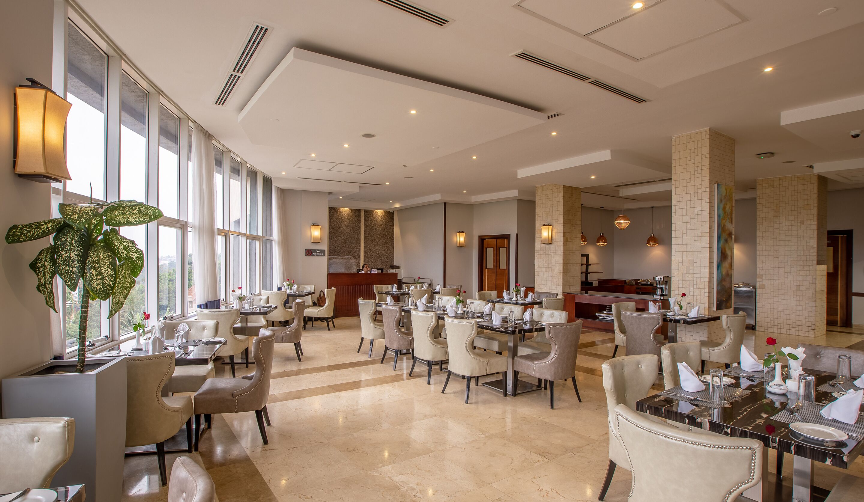 A spacious hotel dining area with multiple tables and chairs, large windows, and a reception desk.