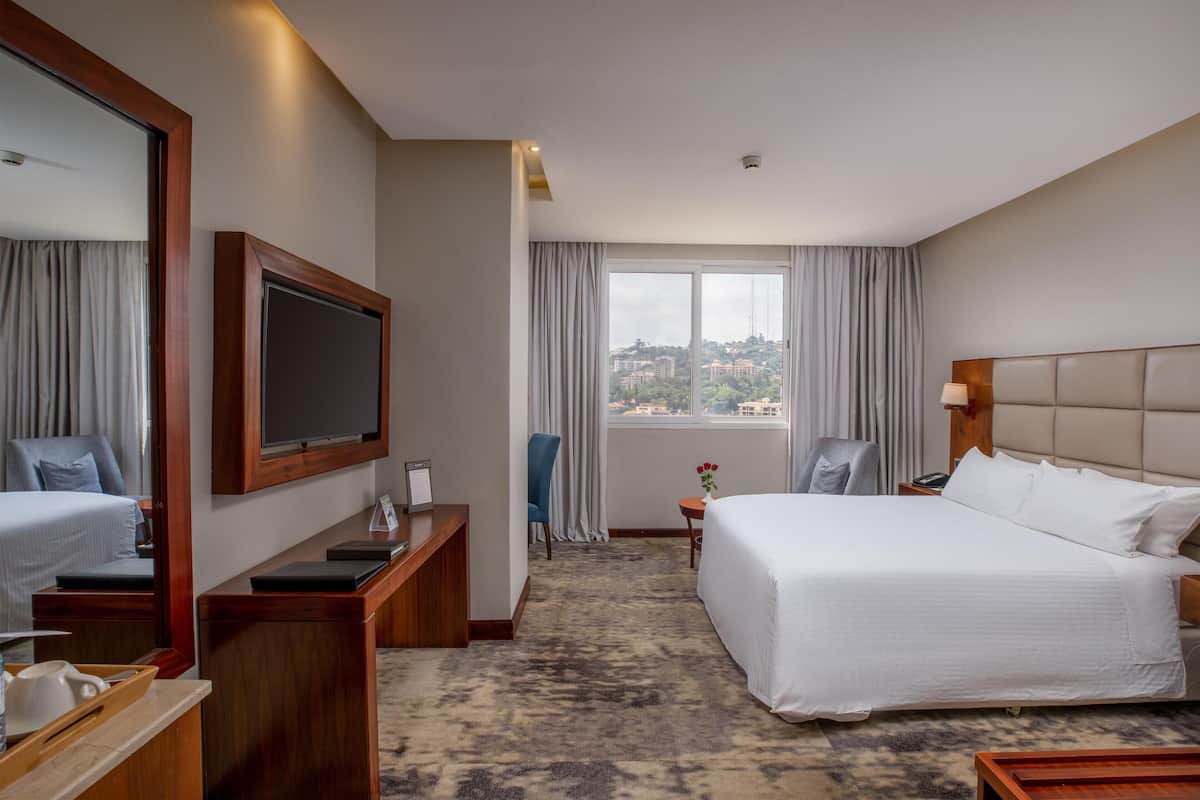 Een hotelkamer met een groot bed, een bureau met een flatscreen-tv, een stoel, een klein tafeltje met een bloevas en uitzicht op een stadsbeeld.