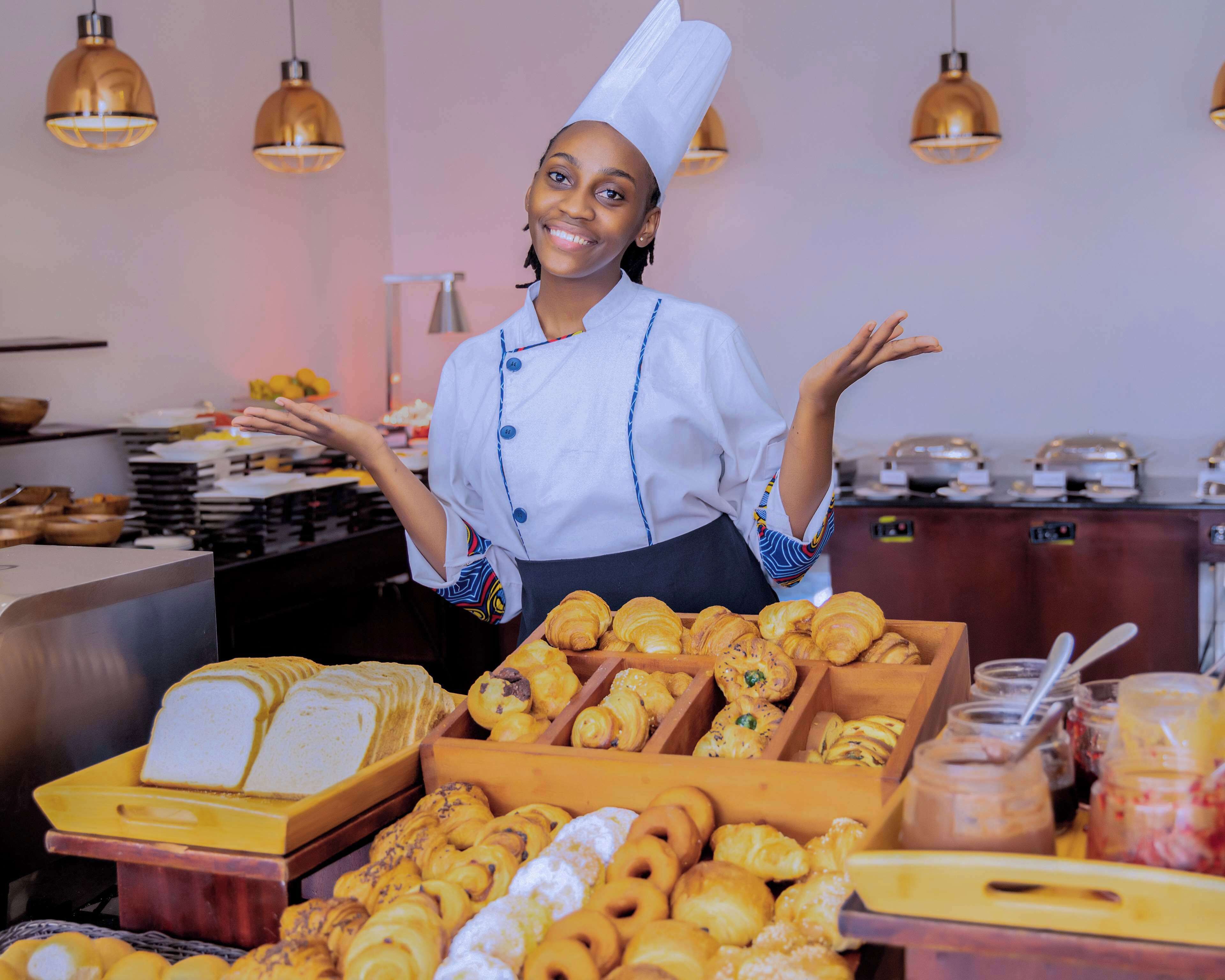 Une personne vêtue d’un uniforme de chef se tient dans une boulangerie, où diverses pâtisseries sont exposées.