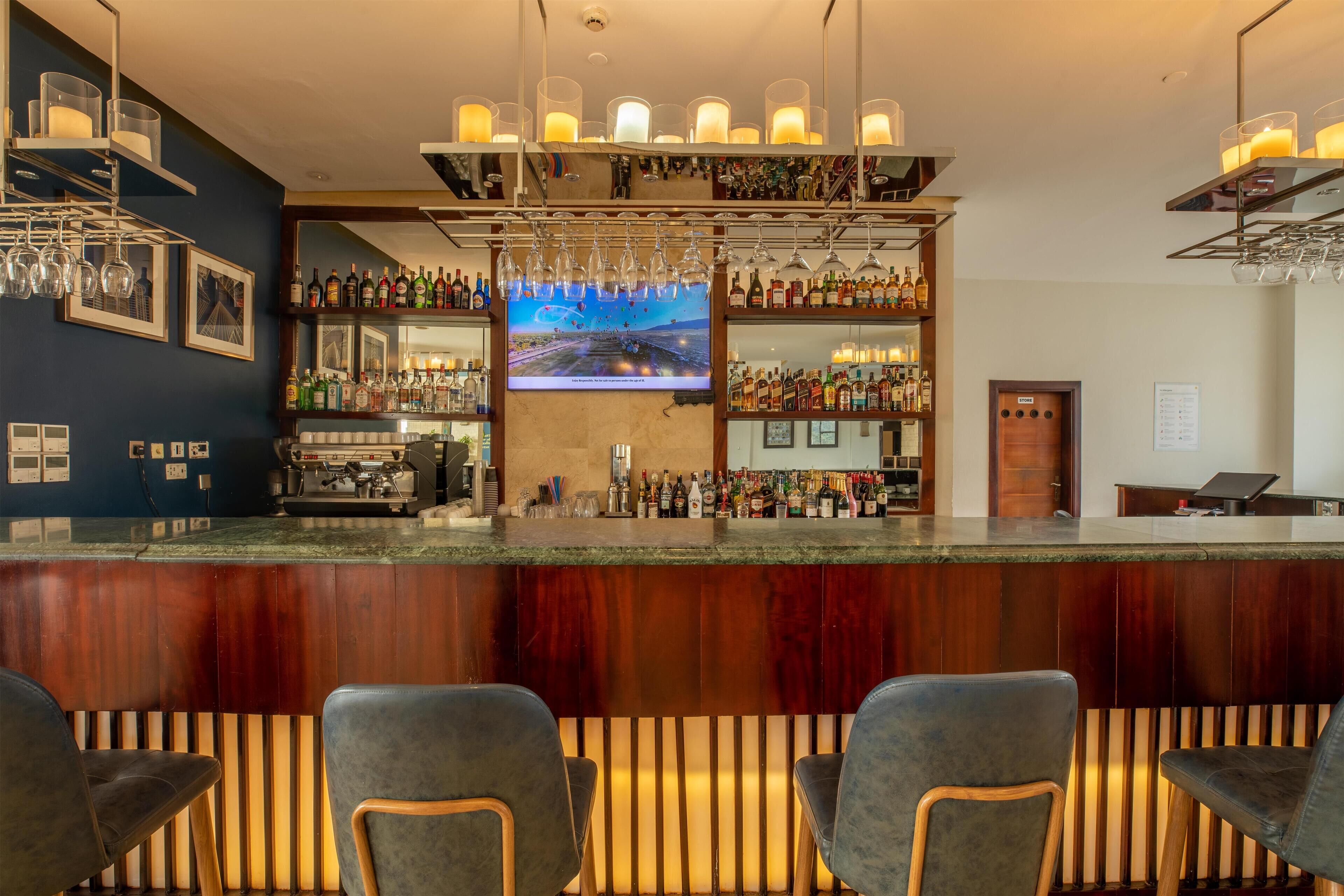 Zona de bar con mostrador de madera, varias estanterías llenas de botellas, vasos colgantes y una pantalla de televisión.