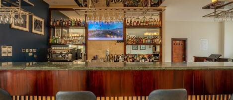 Zona de bar con mostrador de madera, varias estanterías llenas de botellas, vasos colgantes y una pantalla de televisión.