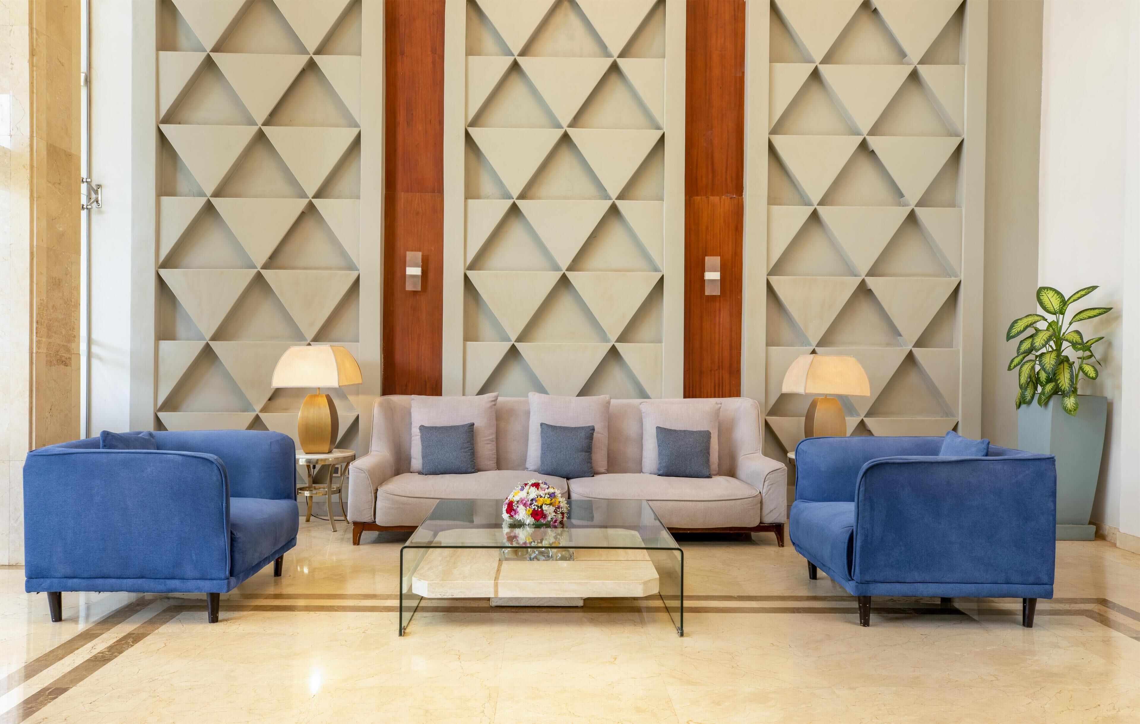 Eine Lobby mit einem beigen Sofa, zwei blauen Sessel, einem Glastisch und einer Wand mit geometrischem Muster.