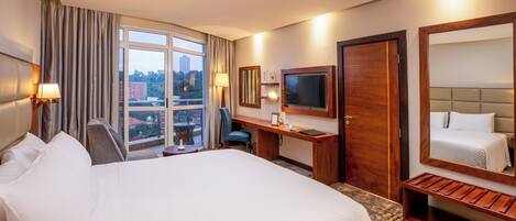 Quarto de hotel com uma cama grande, uma escrivaninha com cadeira, uma televisão e uma janela com cortinas.