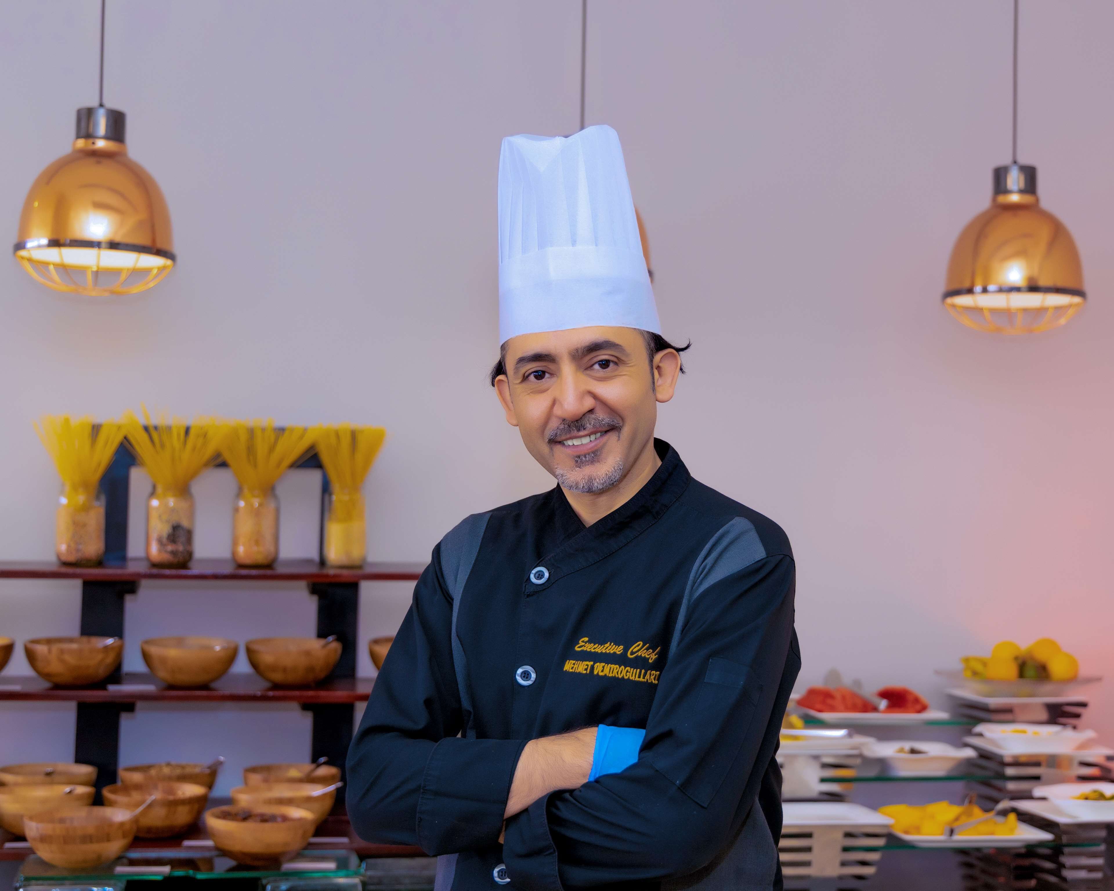 Un chef con uniforme negro y gorro blanco, de pie en una cocina con luces colgantes y diversos utensilios de cocina e ingredientes en estantes.