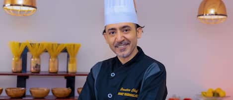 A chef in a black uniform with a white chef's hat, standing in a kitchen with hanging lights and various cooking utensils and ingredients on shelves.