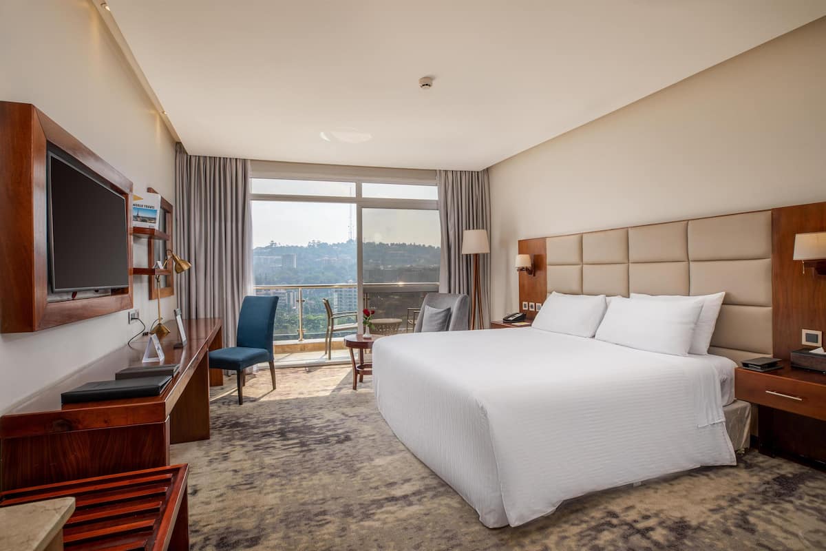 Een hotelkamer met een groot bed, een bureau met een televisie, een stoel, een kleine tafel met een vaas, en een stadsgezicht door grote ramen.