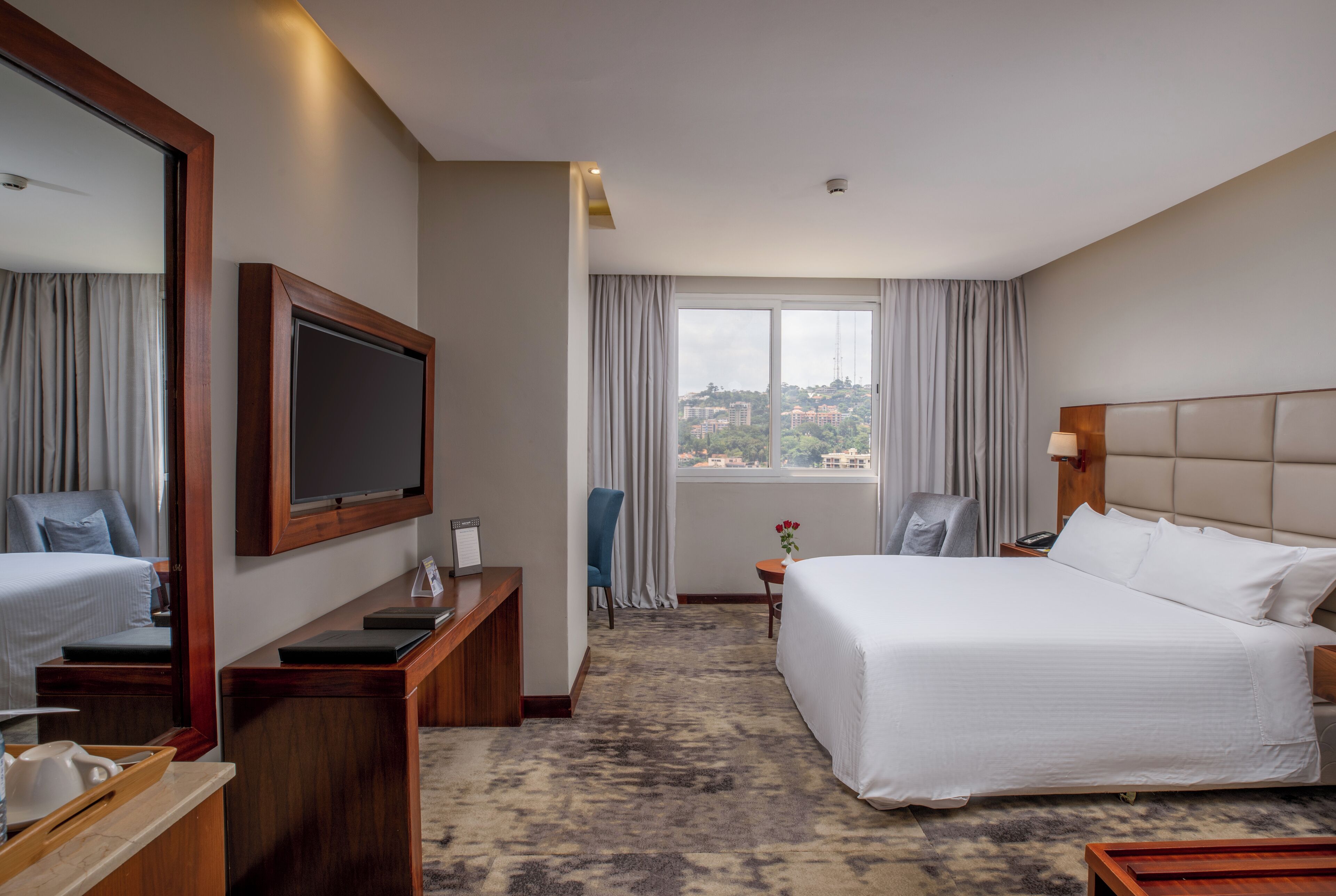 A hotel room with a large bed, a desk with a computer, a TV, and a view of a cityscape through the window.