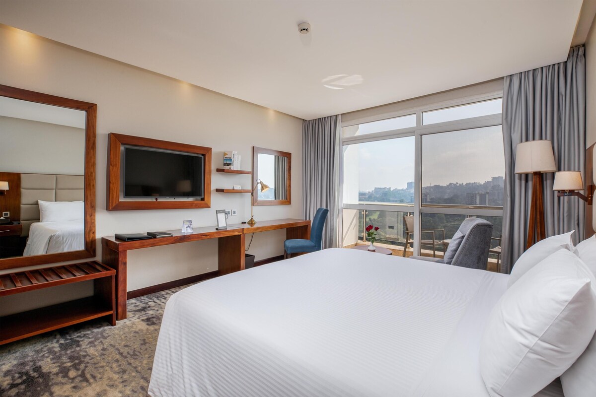 Habitación de hotel con una cama grande, escritorio, silla, televisor y vista a la ciudad.